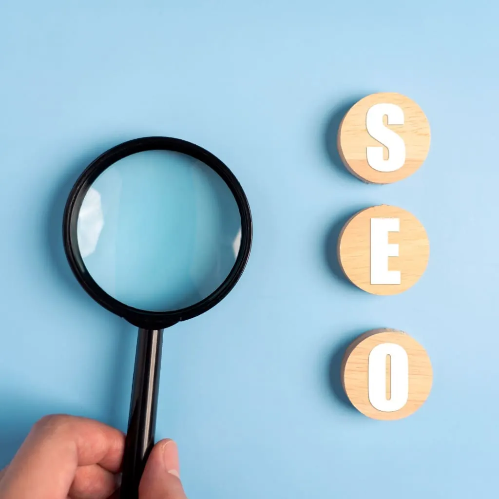 Hand holding a magnifying glass next to wooden discs spelling SEO on a blue background