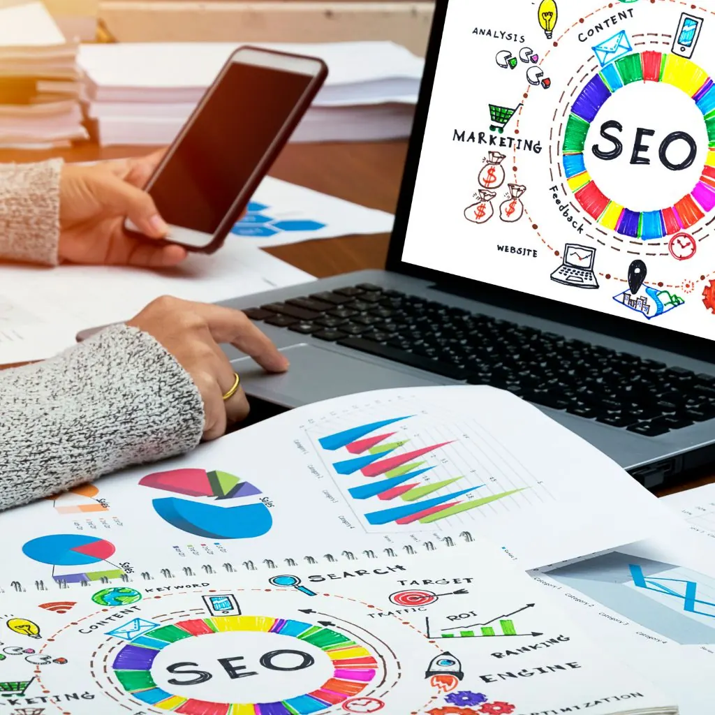 Person using a phone and laptop surrounded by printed SEO and marketing charts